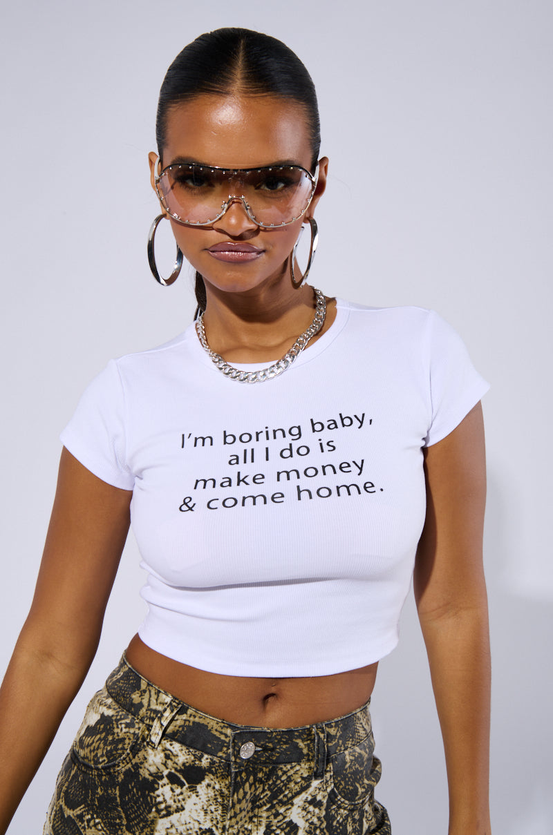 A person wearing sunglasses, hoop earrings, a silver necklace, and an "ALL I DO IS MAKE MONEY GRAPHIC BABY TEE" in a ribbed knit cotton blend. The cropped fit pairs perfectly with their high-waisted patterned pants as they pose against a plain background.
