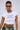 A person wearing sunglasses, hoop earrings, a silver necklace, and an "ALL I DO IS MAKE MONEY GRAPHIC BABY TEE" in a ribbed knit cotton blend. The cropped fit pairs perfectly with their high-waisted patterned pants as they pose against a plain background.