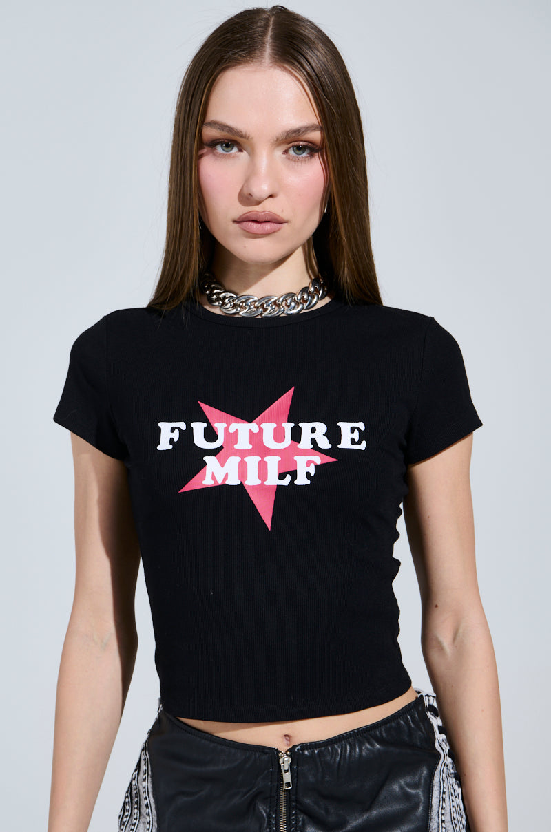 A person with long brown hair wears the FUTURE MILF BABY TEE, featuring the phrase "Future MILF" over a pink star. They accessorize with a chunky silver chain necklace and stand against a plain gray background, exuding effortless style. 0