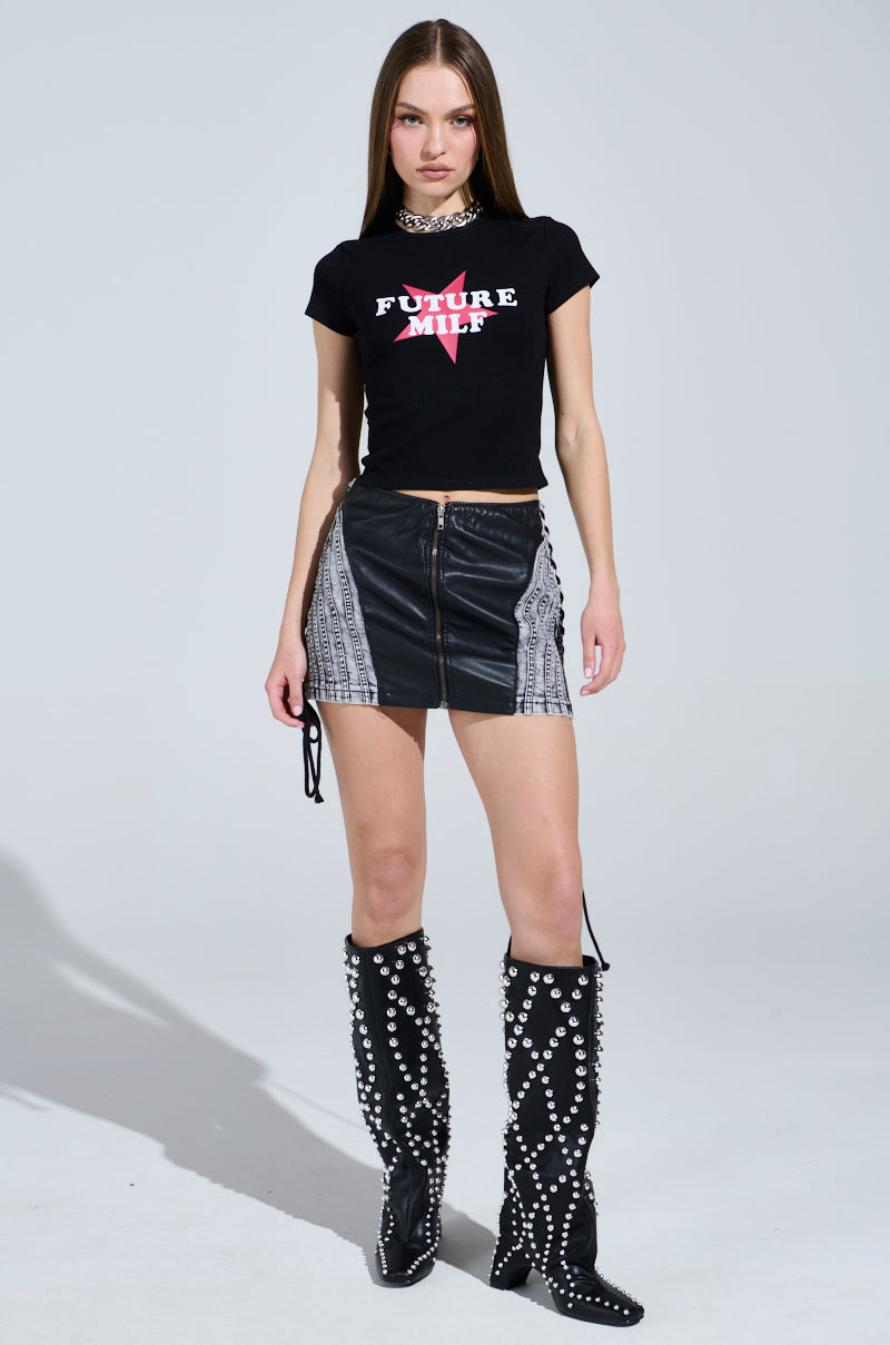 A woman stands confidently in a ribbed knit FUTURE MILF BABY TEE, paired with a black leather mini skirt featuring patterned panels. Her knee-high black boots with pearl-like beads and straight hair complete the ensemble as she poses against a plain background. 4