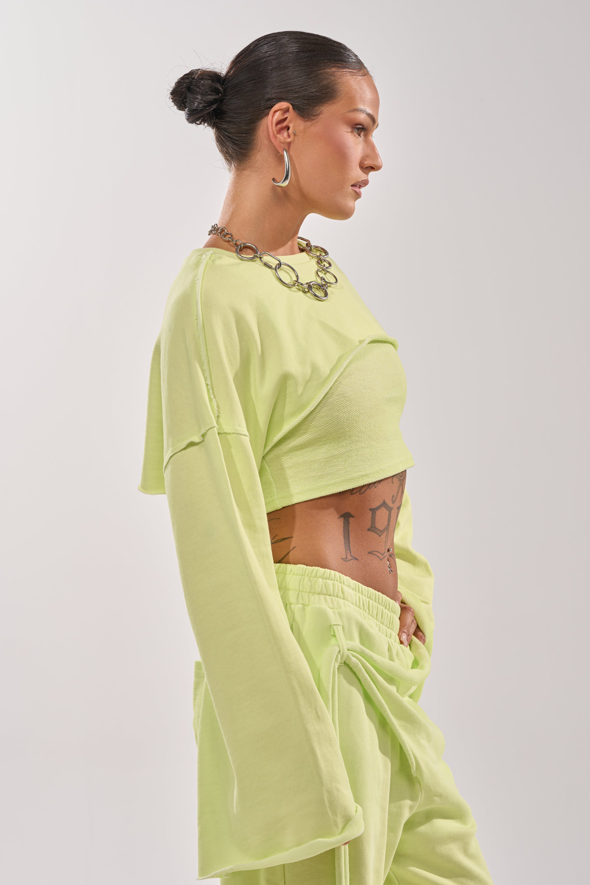 A woman with a bun, large hoops, chunky necklace, and tattoos wears the TRY ME BABY CROP SWEATSHIRT in light green with matching pants, standing in profile against a plain white background. 2