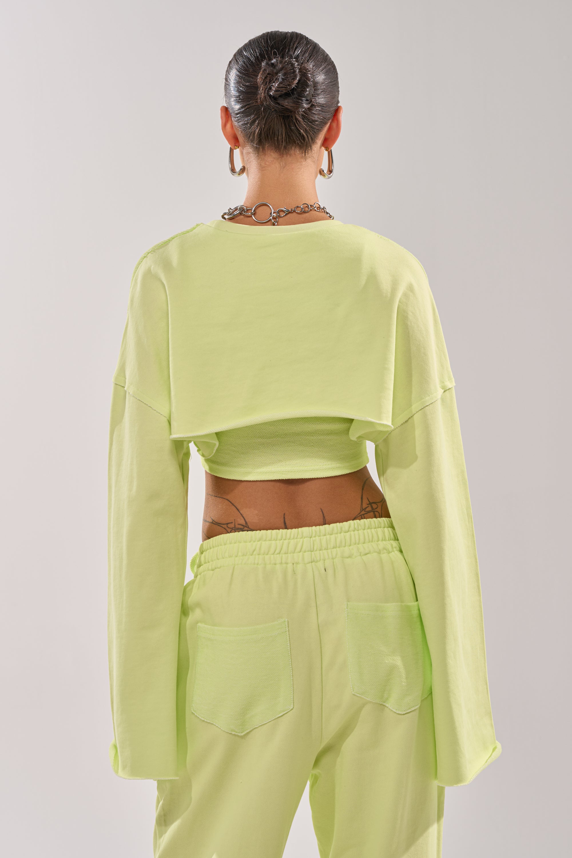 A person with slicked-back hair and hoop earrings is shown from the back, wearing the TRY ME BABY CROP SWEATSHIRT in lime with matching pants featuring large back pockets. Tattoos are partially visible on their lower back and waist. 3