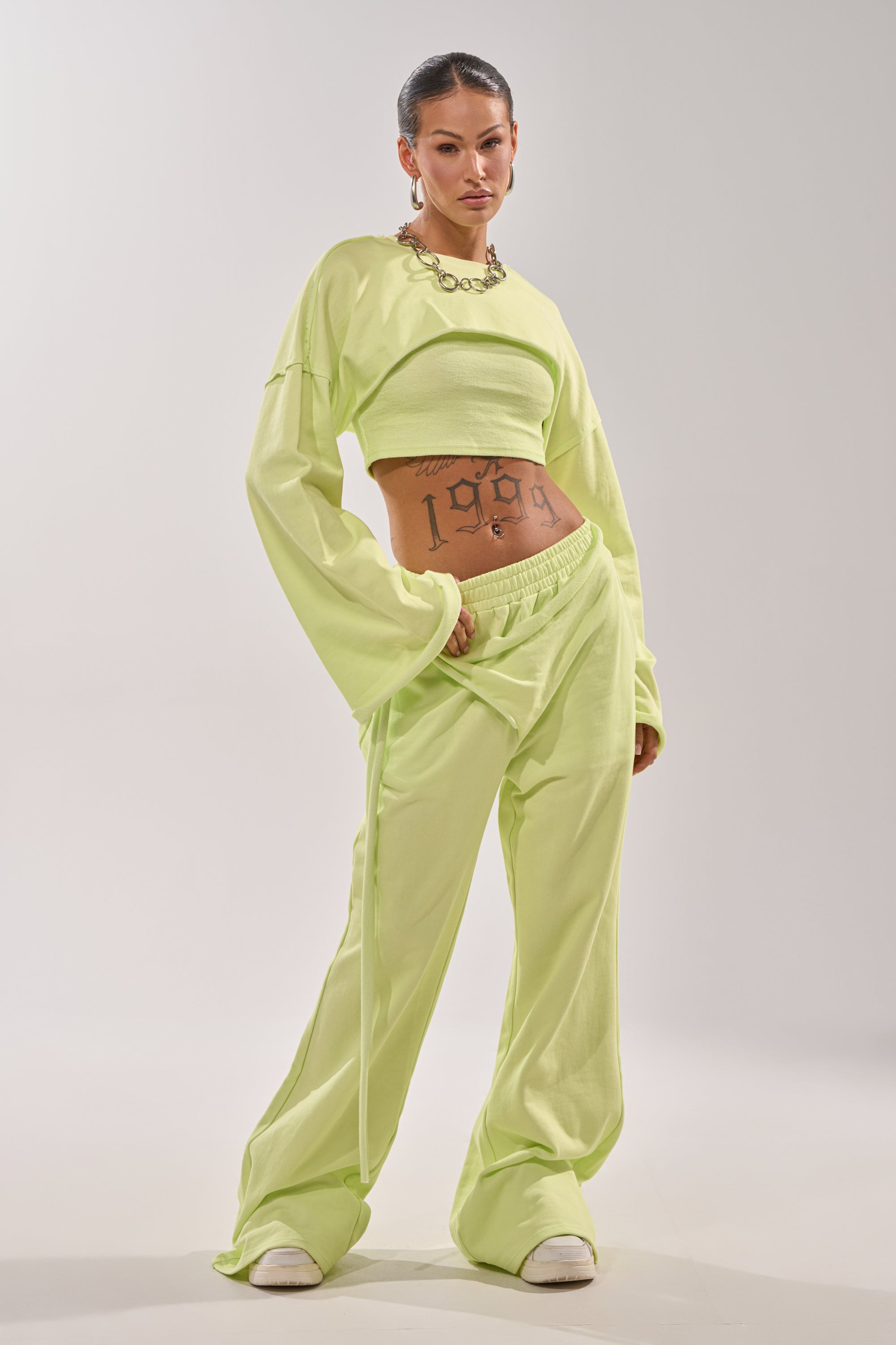 A woman poses confidently in the TRY ME BABY CROP SWEATSHIRT—light green, cropped silhouette with matching wide-leg pants. She pairs it with white sneakers, a chunky chain necklace, and shows off her "1998" midriff tattoo against a white background. 4