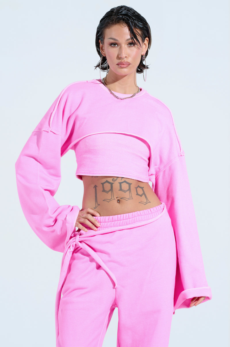 Donning a vibrant pink ISSA DOUBLE LAYER CROP SWEATSHIRT with matching sweatpants, the individual confidently poses with one hand on their hip. The "1999" tattoo on their midriff contrasts against the light blue backdrop, merging style and attitude.