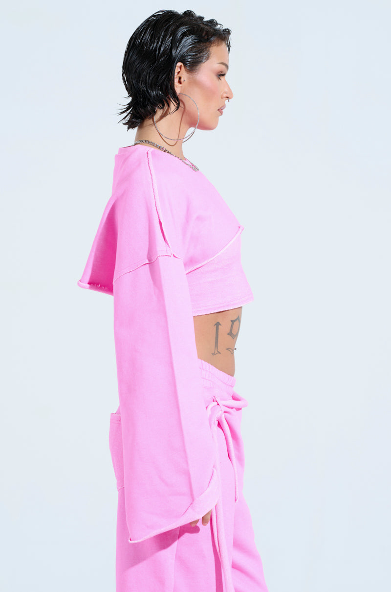 A person with short, dark hair is wearing a loose-fitting, light pink ISSA Double Layer Crop Sweatshirt and matching pants in an athleisure style with wide sleeves. A tattoo is visible on their side, set against a plain, light background.