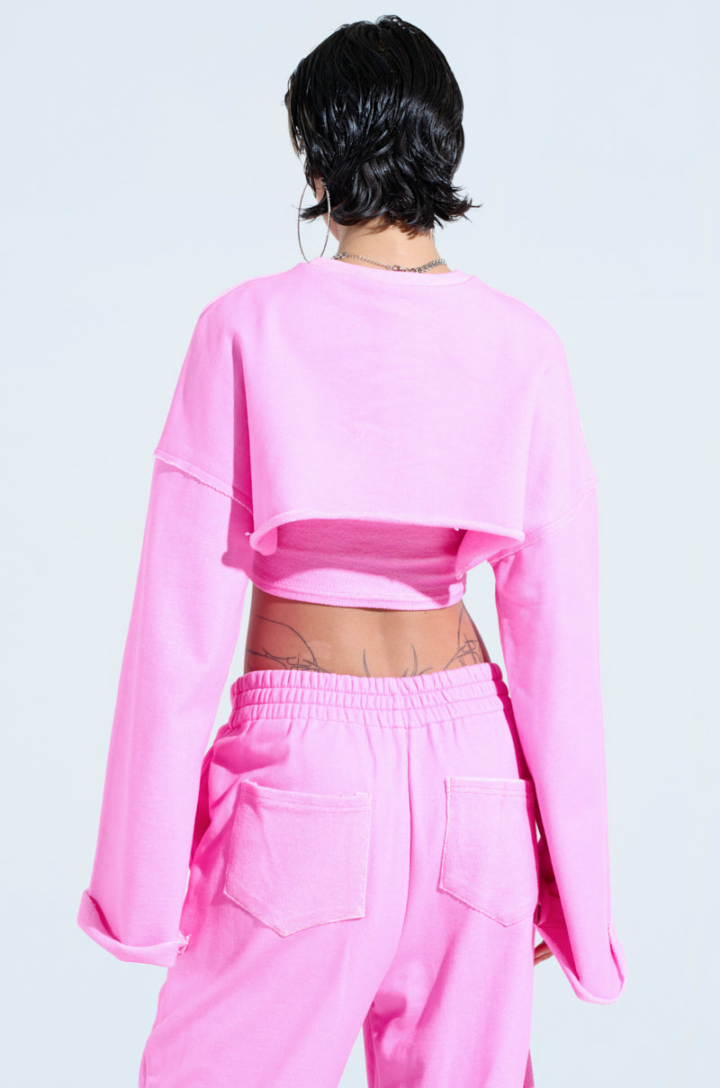 A person with short black hair is seen from behind, wearing an oversized ISSA DOUBLE LAYER CROP SWEATSHIRT in pink paired with matching pants. Tattoos adorn their lower back, and large hoop earrings add flair as they stand against a light blue background, embodying the perfect athleisure ensemble.