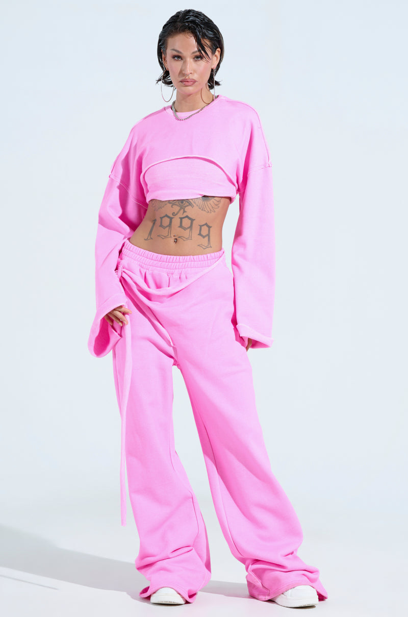 A person wears a bright pink athleisure set, featuring the ISSA DOUBLE LAYER CROP SWEATSHIRT and wide-leg pants. Their dark slicked-back hair and midriff tattoos complement their style. White shoes peek out, while a light-colored background enhances the outfit's oversized silhouette.