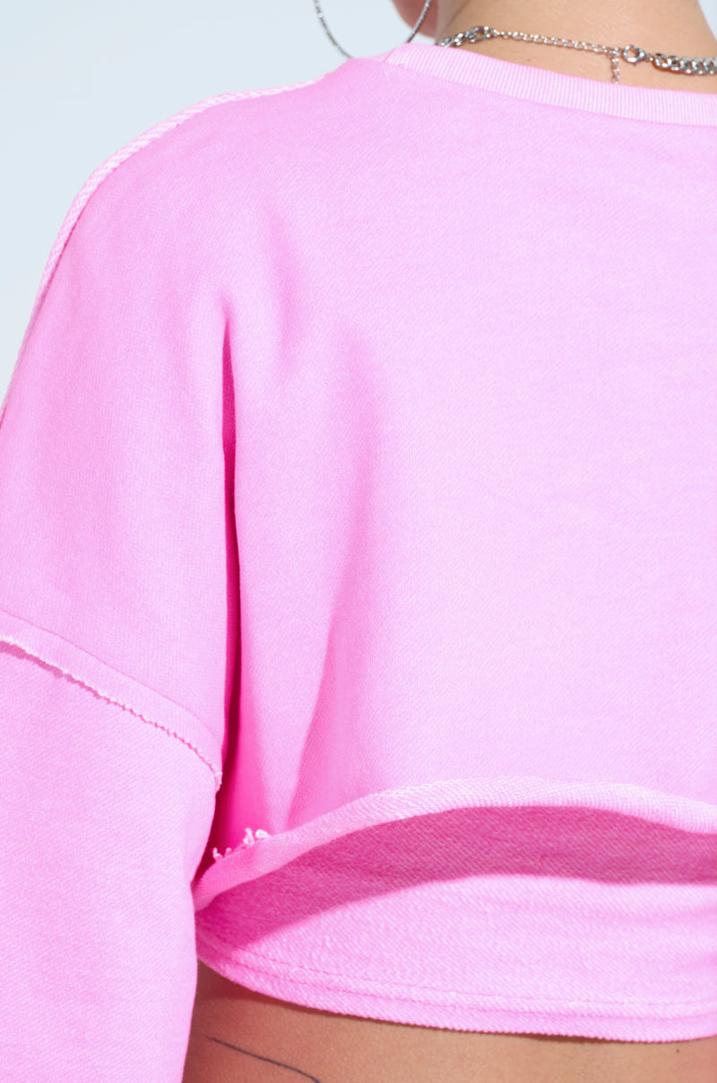 A person wearing the ISSA DOUBLE LAYER CROP SWEATSHIRT in bright pink, showcasing visible stitching, stands with their back to the camera. They accessorize with a hoop earring and a delicate chain necklace, completing their athleisure look against a calming light blue backdrop.