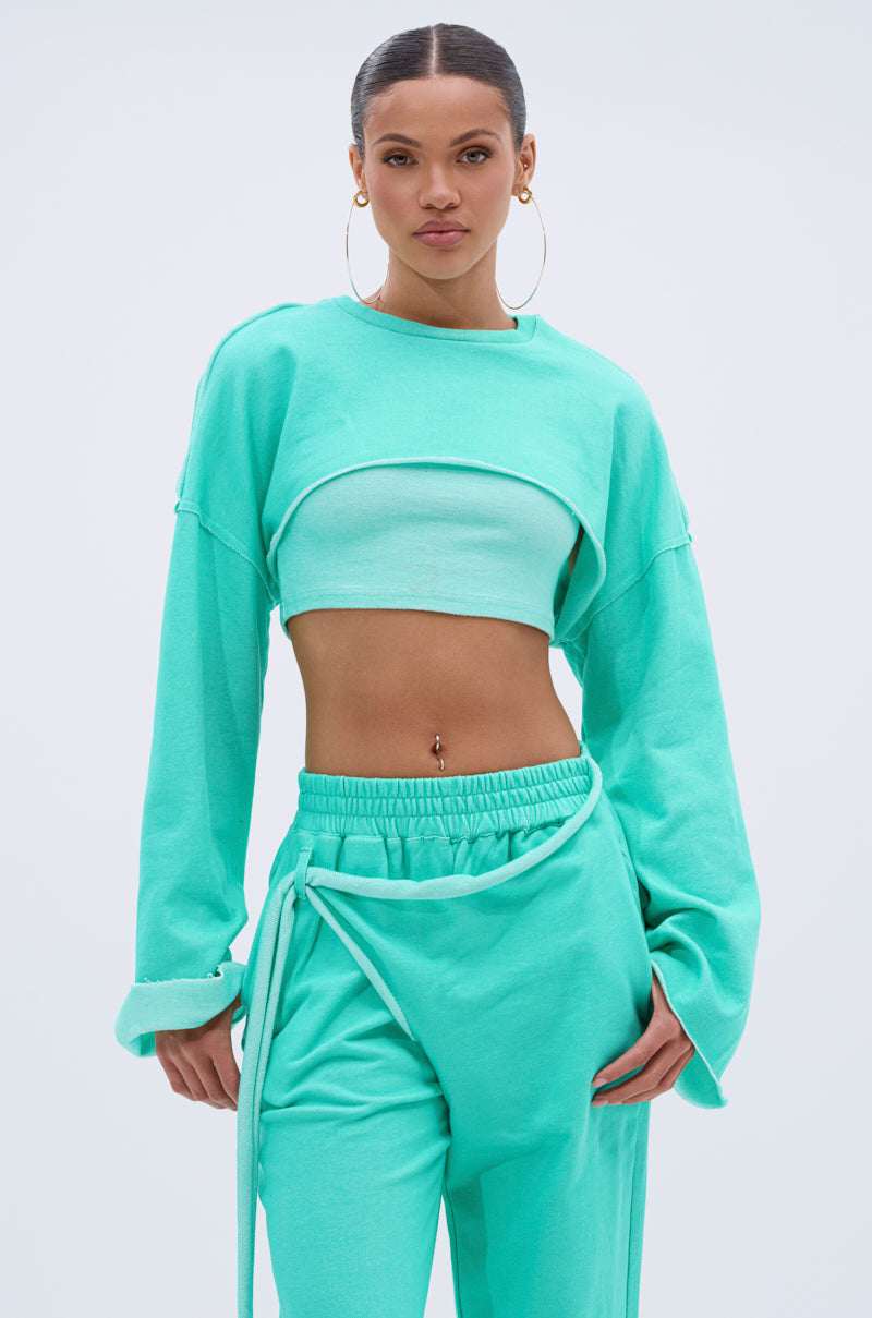 A woman wears the ISSA DOUBLE LAYER CROP SWEATSHIRT IN TIFFANY over a matching sports bra and loose high-waisted pants, accessorized with hoop earrings and a belly button piercing, standing against a plain light blue background. 0