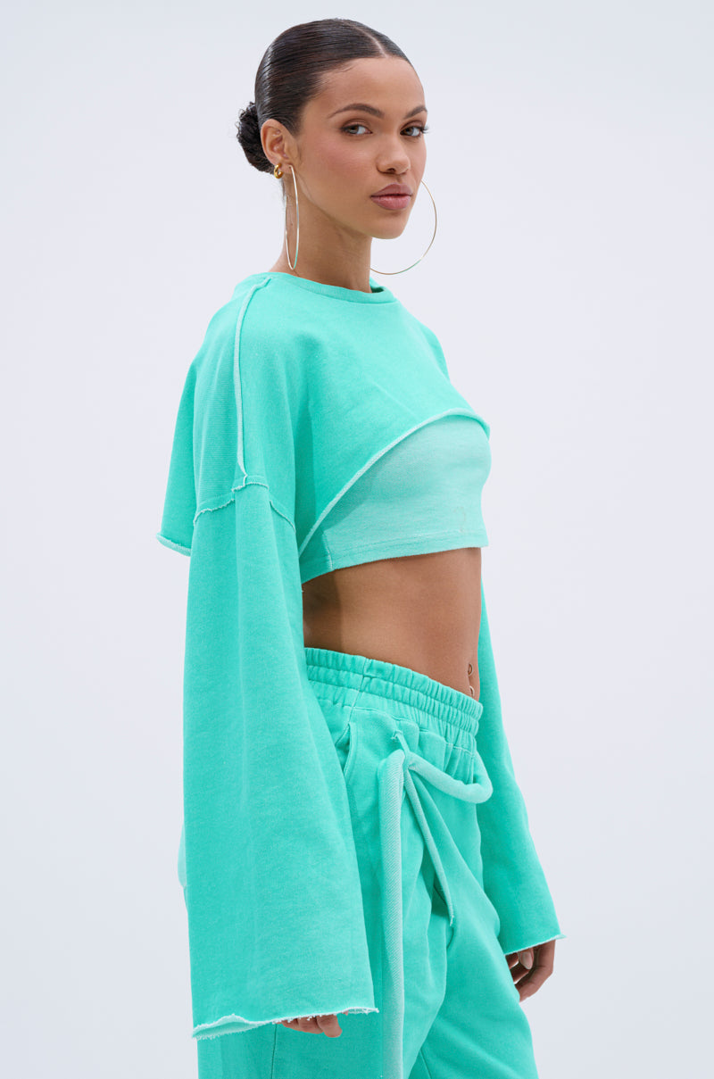 A woman stands against a plain light background wearing the ISSA DOUBLE LAYER CROP SWEATSHIRT IN TIFFANY by AKIRA Label, styled with wide sleeves, hoop earrings, and her hair in a neat bun. 2