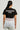 A person with long hair is wearing a black AKIRA Label EVERYBODYS TYPE SHIRT featuring white text. They have a white lace garment over light-colored pants and stand against a neutral background.