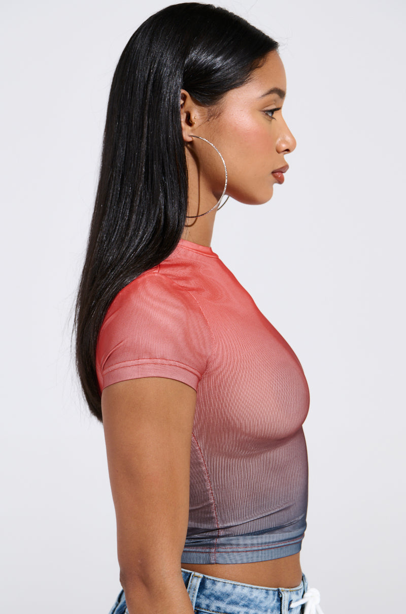 A woman with long dark hair and large hoop earrings wears the snug-fit DID IT AGAIN OMBRE MESH TSHIRT by AKIRA, which transitions from red to blue. She faces sideways, showcasing her profile against a plain backdrop.