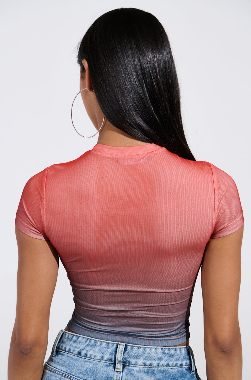A woman with long black hair faces away, wearing a DID IT AGAIN OMBRE MESH TSHIRT that fades from dark gray to red. Large hoop earrings and light blue jeans complement the snug fit of her cropped top against a plain white backdrop.