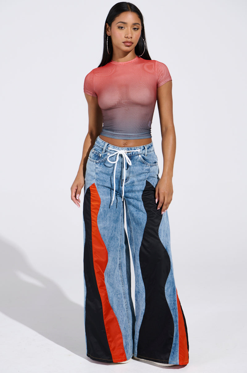 A woman stands against a plain background wearing an AKIRA Label DID IT AGAIN OMBRE MESH T-SHIRT with a fitted cropped design and wide-legged jeans in red, black, and blue panels. Her long dark hair flows as she confidently poses with her hands by her sides.