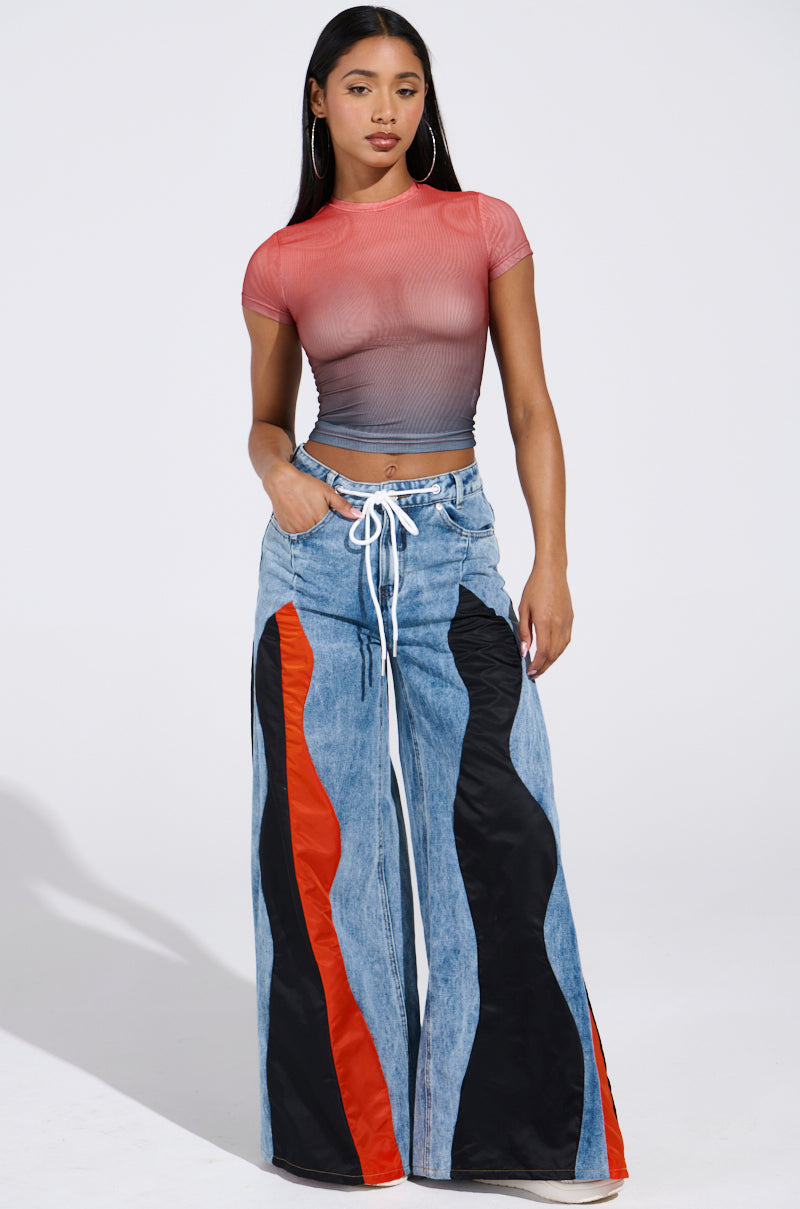 Wearing the DID IT AGAIN OMBRE MESH TSHIRT transitioning from red to gray, paired with high-waisted wide-leg jeans in bold orange and black wavy patterns, a woman stands confidently. Her long dark hair cascades down, framed by large hoop earrings as she rests her hands in her jean pockets.
