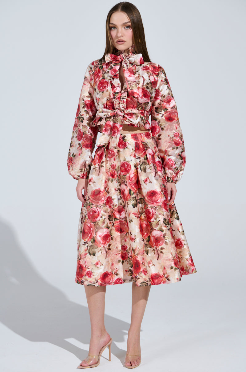 A woman models a vintage-inspired outfit featuring the "Dance With Me Floral Midi Skirt" with long sleeves, high collar, and rose pattern. The knee-length skirt accompanies clear high-heeled shoes against a plain light-colored background.