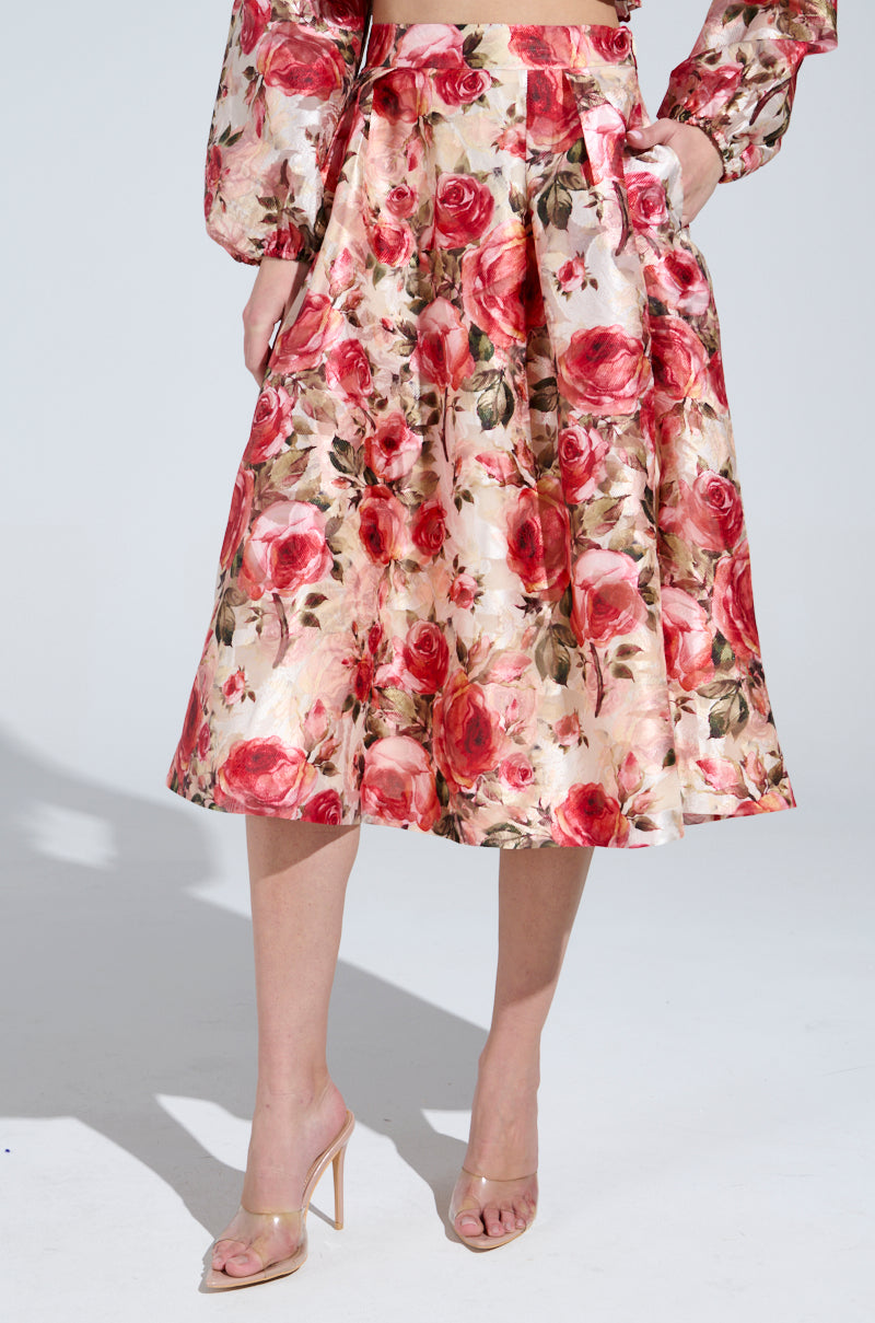 Against a light backdrop, a person in vintage-inspired attire features the "DANCE WITH ME FLORAL MIDI SKIRT," paired with a rose-patterned top. With one hand in their skirt pocket, they elegantly wear nude high-heeled sandals.
