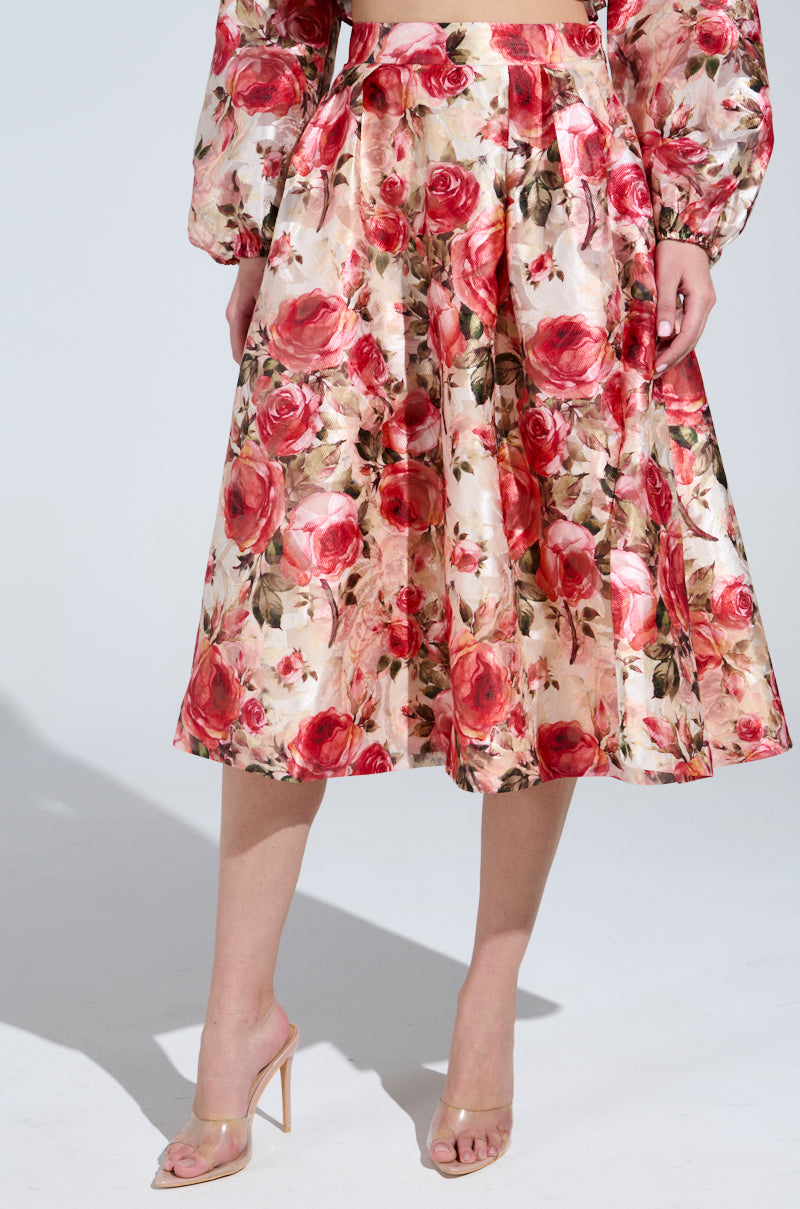 A person in a vintage-inspired look features the DANCE WITH ME FLORAL MIDI SKIRT with large red and pink roses and green leaves on a light background, paired with a long-sleeved top and stylish clear high-heeled sandals.