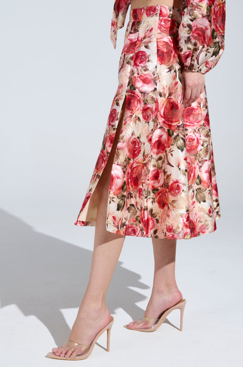 Wearing the "DANCE WITH ME FLORAL MIDI SKIRT," adorned with vintage-inspired red roses and a high slit that reveals the left leg, they pair it with a matching top and clear heeled sandals against a plain white backdrop.