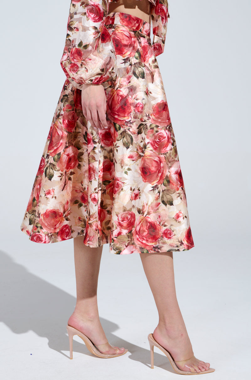 A person confidently walks in the DANCE WITH ME FLORAL MIDI SKIRT featuring rose patterns with red and pink blooms, paired with neutral high-heeled sandals. Only their lower body is visible against a softly shaded background.