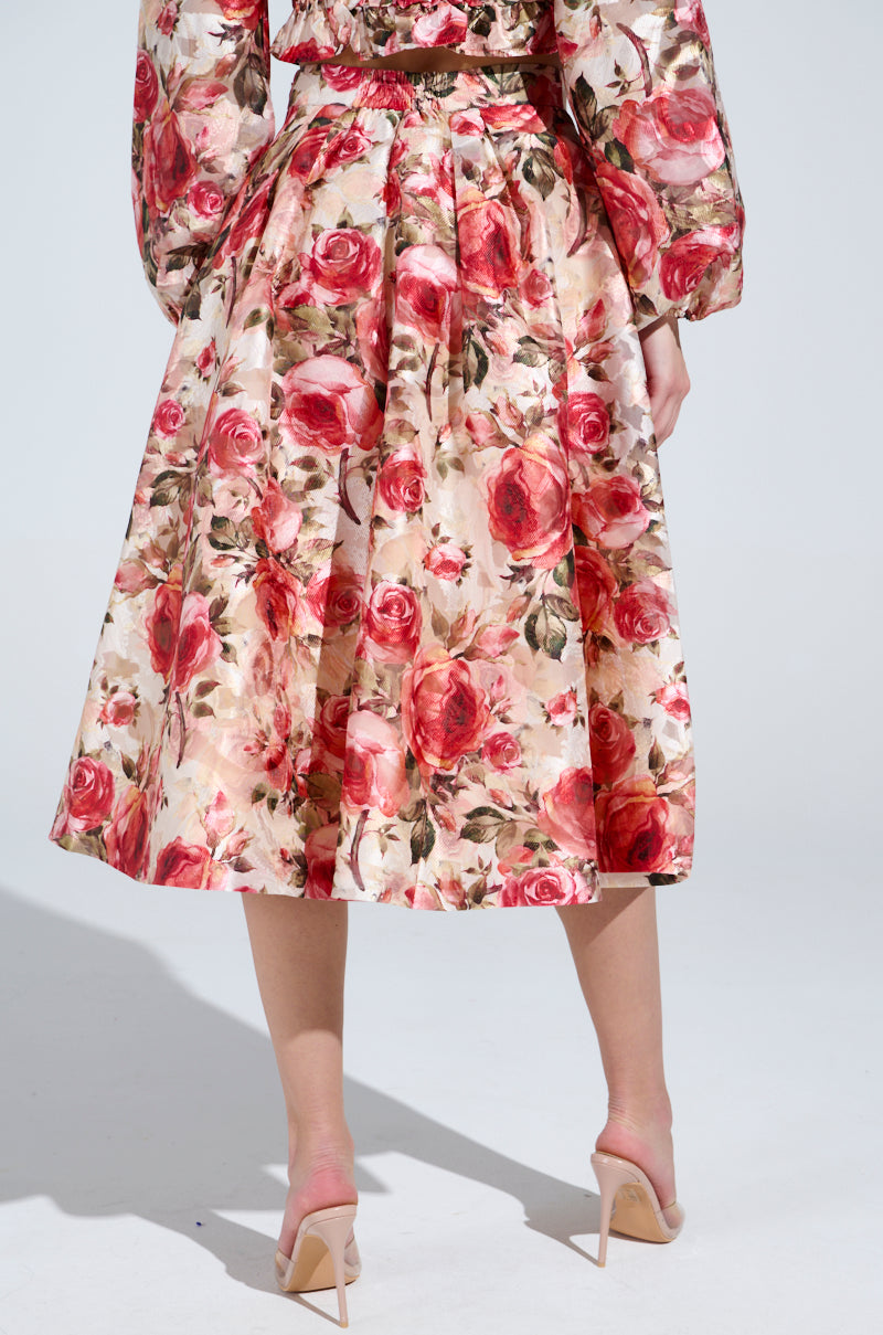 A person wearing the DANCE WITH ME FLORAL MIDI SKIRT with a vintage-inspired rose pattern and long sleeves stands back to the camera, exuding timeless elegance. They pair it with nude high-heeled shoes against a plain white backdrop.