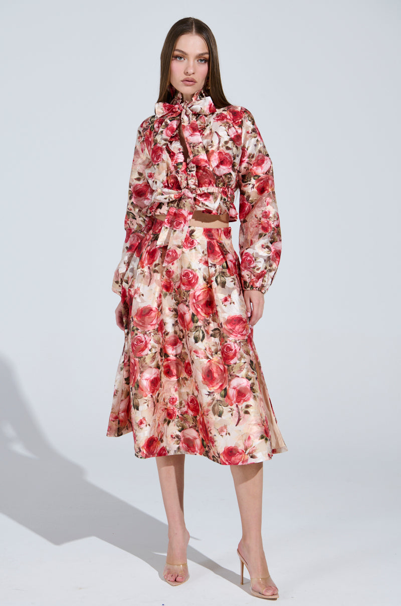 A woman wears a Dance With Me Blouse and the DANCE WITH ME FLORAL MIDI SKIRT, featuring a vintage rose pattern in red and pink. She stands against a plain gray background, enhancing her look with transparent heels.