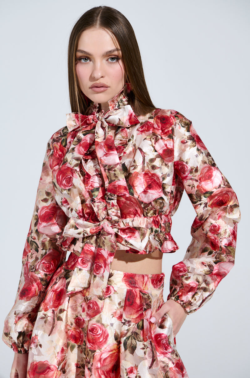 A woman poses against a plain background in the DANCE WITH ME FLORAL TIE FRONT BLOUSE and matching pants, featuring an elegant rose pattern. The blouse cinches at the waist and ties at the neck. Her long brown hair flows loosely, with a neutral expression on her face.
