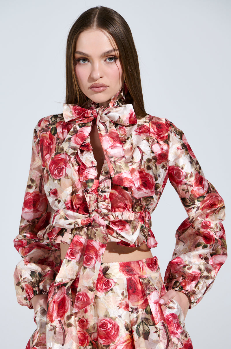 A person with long brown hair wears a vintage-inspired outfit showcasing a rose pattern in red and pink hues, featuring the "Dance With Me" floral tie-front blouse with ruffles and matching pants, set against a plain background.