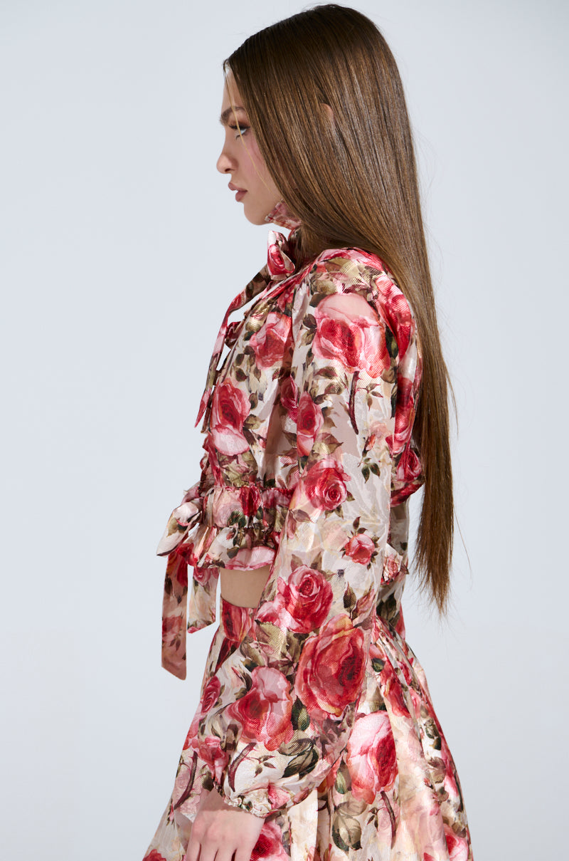 Side view of a person with long brown hair in a vintage-style ensemble featuring the "Dance With Me" floral tie-front blouse adorned with a bow, paired with a flowing skirt, set against a plain gray backdrop.