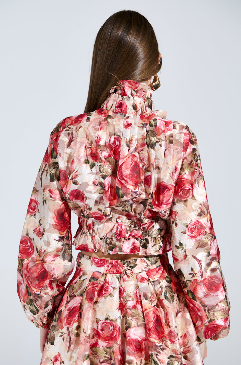 A person with long brown hair wears the "Dance With Me Floral Tie Front Blouse," adorned with vintage-inspired large pink roses and green leaves. It features a high collar, long sleeves, and a pleated waist as they stand facing away from the camera against a plain background.