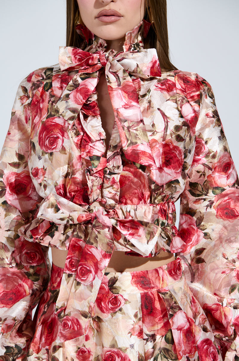 Someone is dressed in a vintage-inspired floral outfit featuring the "Dance With Me Floral Tie Front Blouse," with red and pink rose patterns, chic ruffled details, puffy shoulders, and a matching skirt against a plain light-colored background.