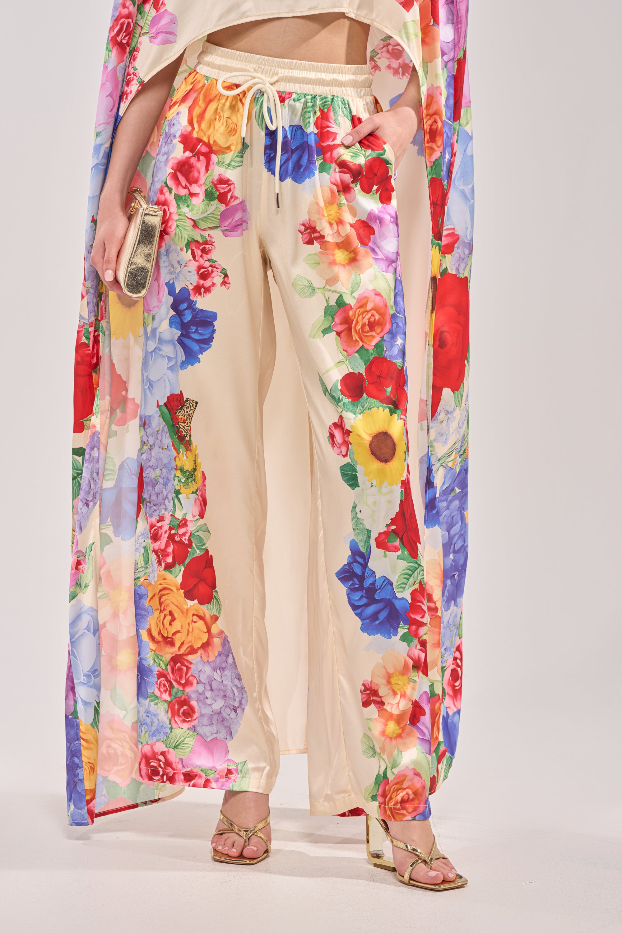 A person wears the FLORAL BURST PALAZZO PANT with a high-rise elastic waistband, paired with a matching floral robe, gold strappy heels, and holds a gold clutch against a plain light-colored background.