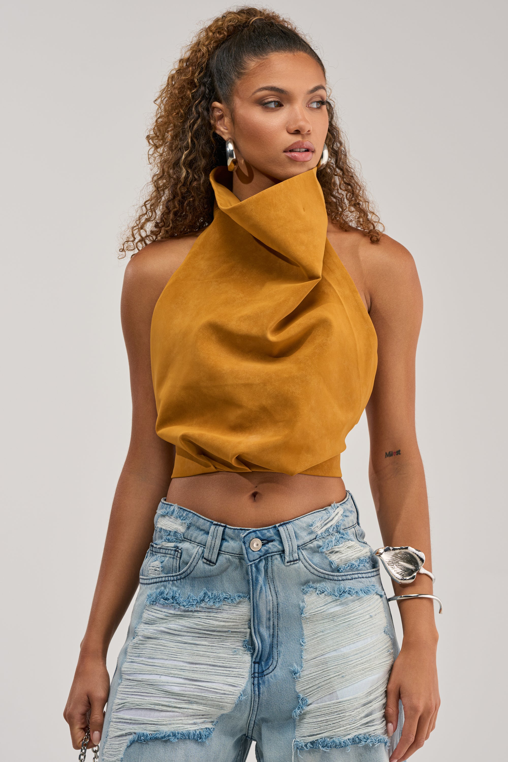 A woman with curly hair in a ponytail wears the JANE SUEDE HALTER TOP IN MUSTARD and light blue distressed high-waisted jeans, accessorized with large hoop earrings, a bracelet, and a visible tattoo on her left forearm.