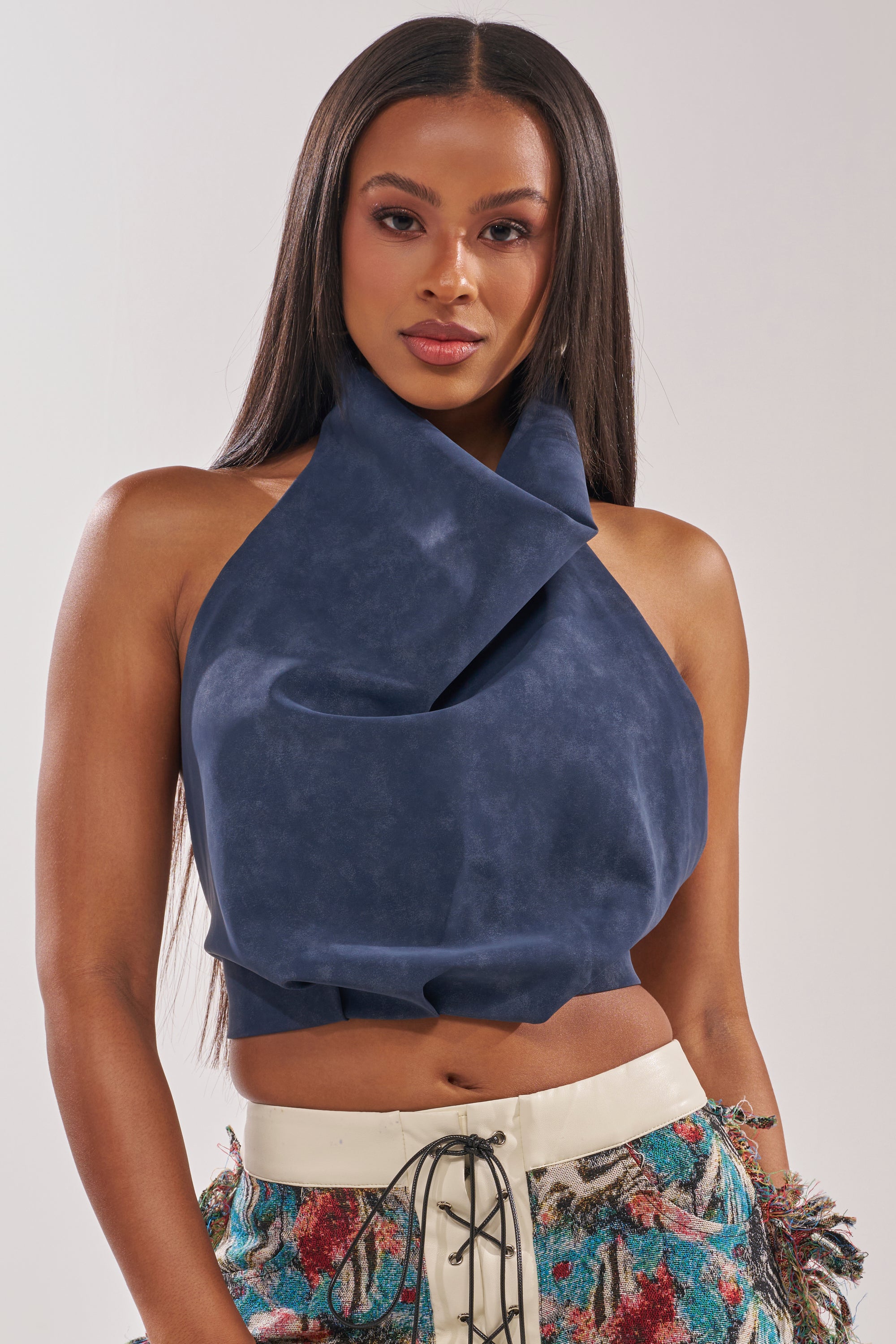 A woman with long straight hair wears the JANE SUEDE HALTER TOP, a sleeveless dark blue halter with a draped front, paired with high-waisted, embroidered lace-up pants. She stands against a plain background, looking at the camera. 0