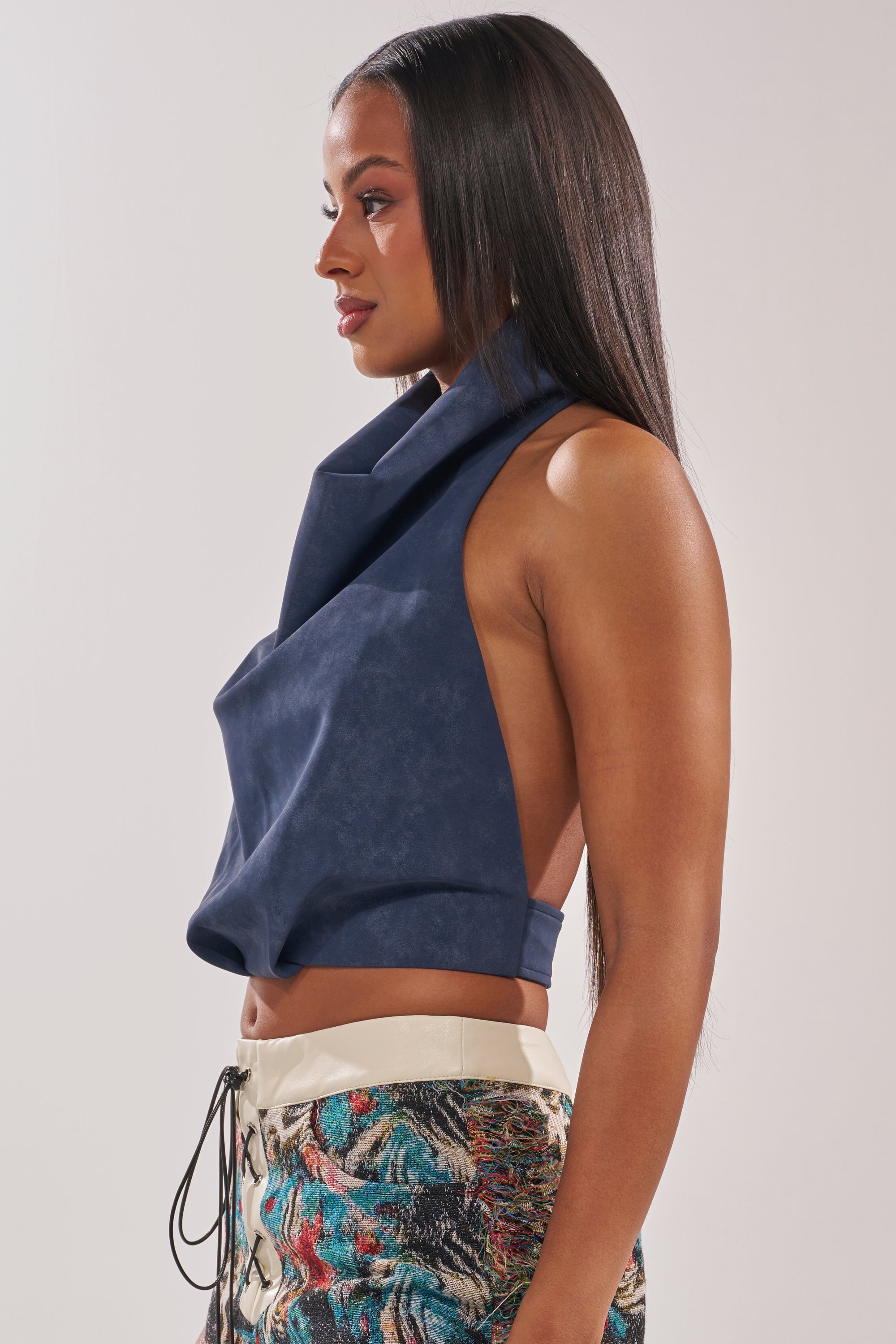 A woman in profile wears the JANE SUEDE HALTER TOP, a sleeveless, open-back navy halter top, paired with colorful abstract-patterned drawstring pants. She has long straight hair and a neutral expression. 2