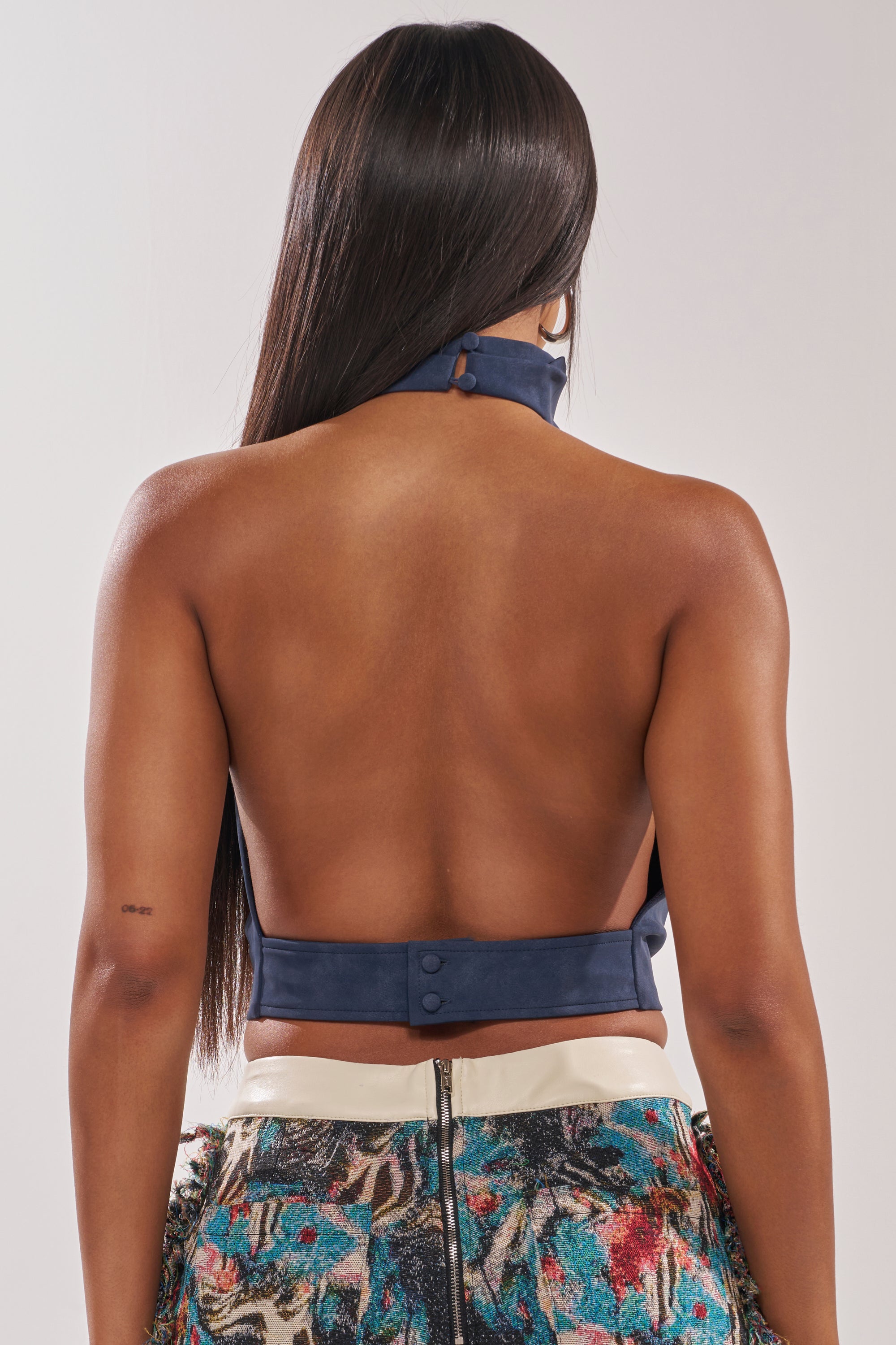 A person with long, straight hair is seen from the back, wearing the JANE SUEDE HALTER TOP in navy with an open-back design and a vibrant patterned skirt featuring a visible zipper. 3