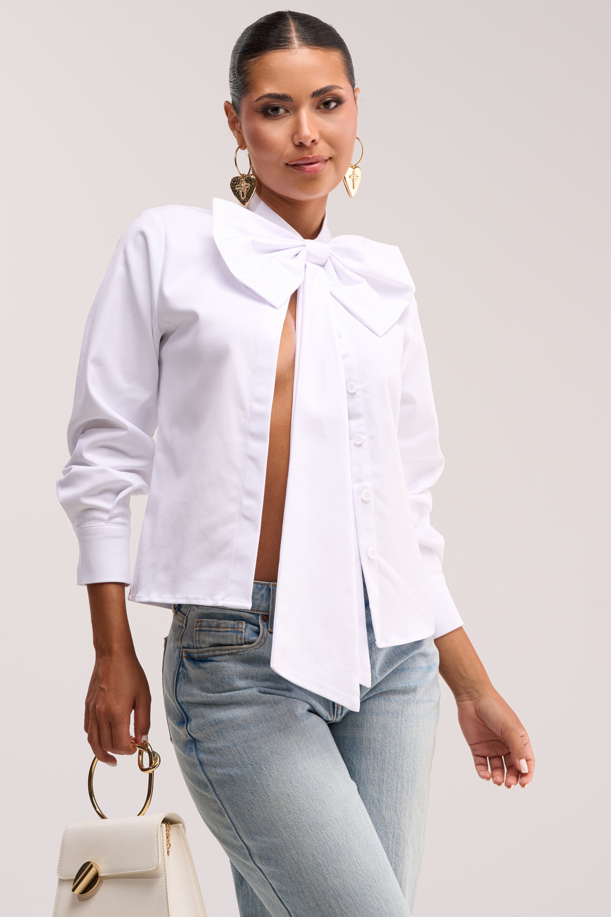 A woman stands against a plain light background wearing the BUSINESS OR PLEASURE POPLIN BLOUSE, featuring an oversized bow at the collar, paired with light blue jeans, gold heart-shaped earrings, and holding a white handbag with a gold handle.