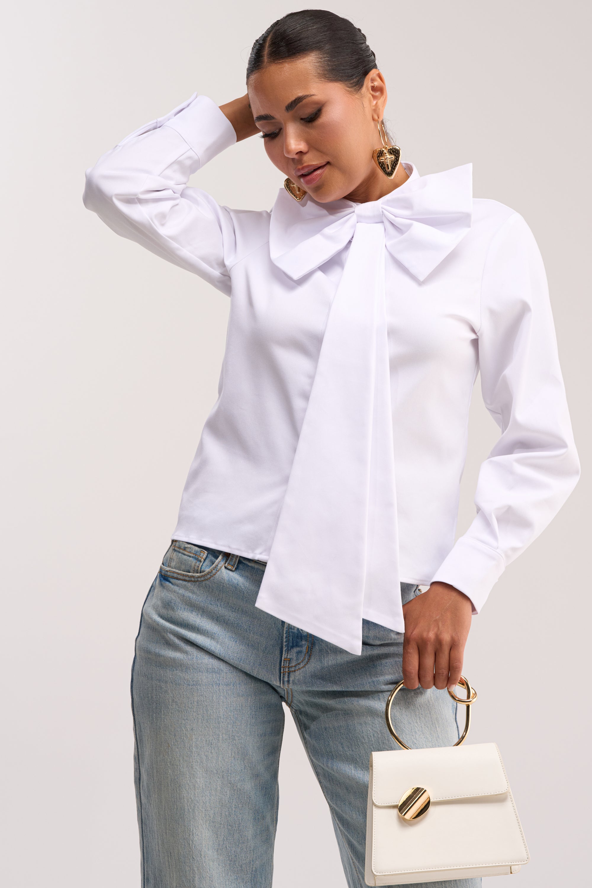 A woman models the BUSINESS OR PLEASURE POPLIN BLOUSE, a modern white poplin top with an oversized bow collar, styled with light blue jeans, gold earrings, and a small white handbag featuring a gold handle. She poses gracefully, hand to head, looking down.