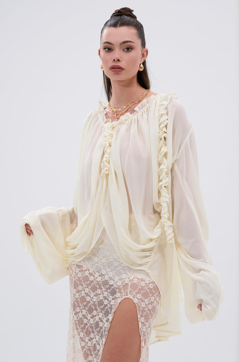 A woman poses against a plain backdrop wearing the LILA WOVEN DRAPEY BLOUSE with ruffled trim and wide sleeves, paired with a lace skirt and gold jewelry. Her sleek bun completes the feminine, flowy silhouette.