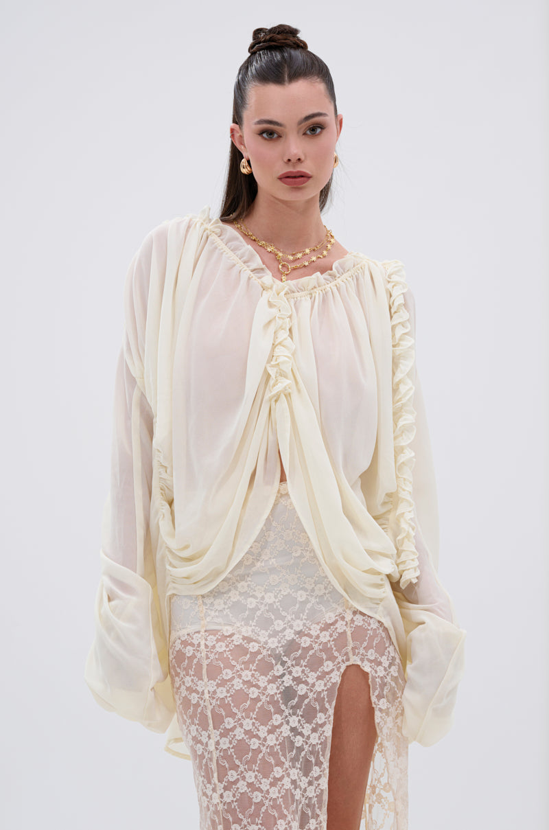 A woman with long dark hair in a high ponytail wears the flowy LILA WOVEN DRAPEY BLOUSE and a cream lace skirt with a high slit. She accessorizes with gold jewelry, creating a feminine look against a plain light background.