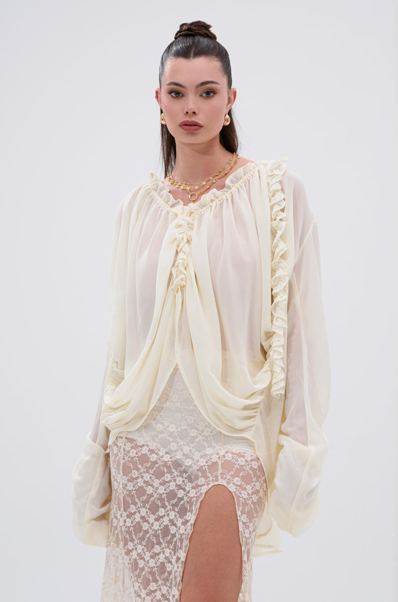 A woman with long dark hair in a high bun wears the LILA WOVEN DRAPEY BLOUSE, paired with a long lace skirt featuring a high slit. Gold earrings and layered necklaces add to her feminine look against a plain light background.