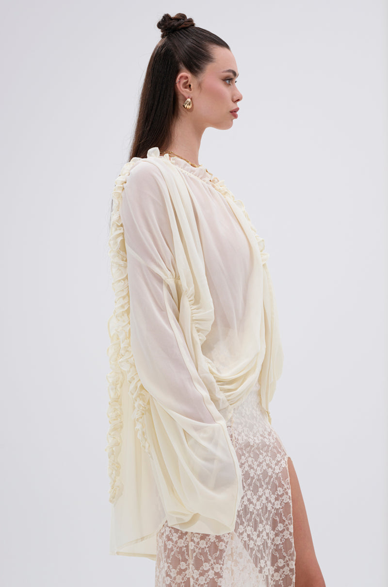 A woman with long dark hair styled half-up poses in profile against a plain light background, wearing the LILA WOVEN DRAPEY BLOUSE with ruffled edges and a white lace skirt with a high slit, creating a feminine vibe.