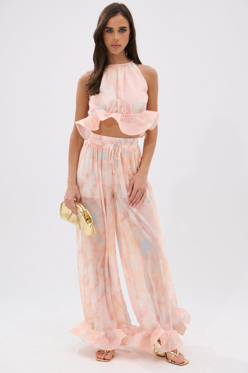 A woman stands against a plain background wearing the COLORFUL SKY OMBRE PALAZZO PANT with ruffle hems and a matching pastel tie-dye sleeveless crop top, paired with gold sandals and a gold clutch. 0