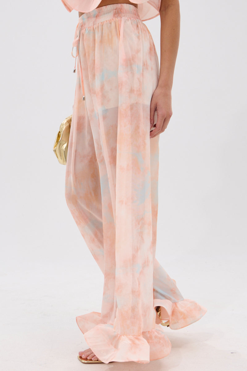 Only the lower half of a person is visible, showcasing the COLORFUL SKY OMBRE PALAZZO PANT—high-rise, wide-leg pants with ruffled hems in peach and light blue—paired with a gold clutch, set against a plain white background. 3