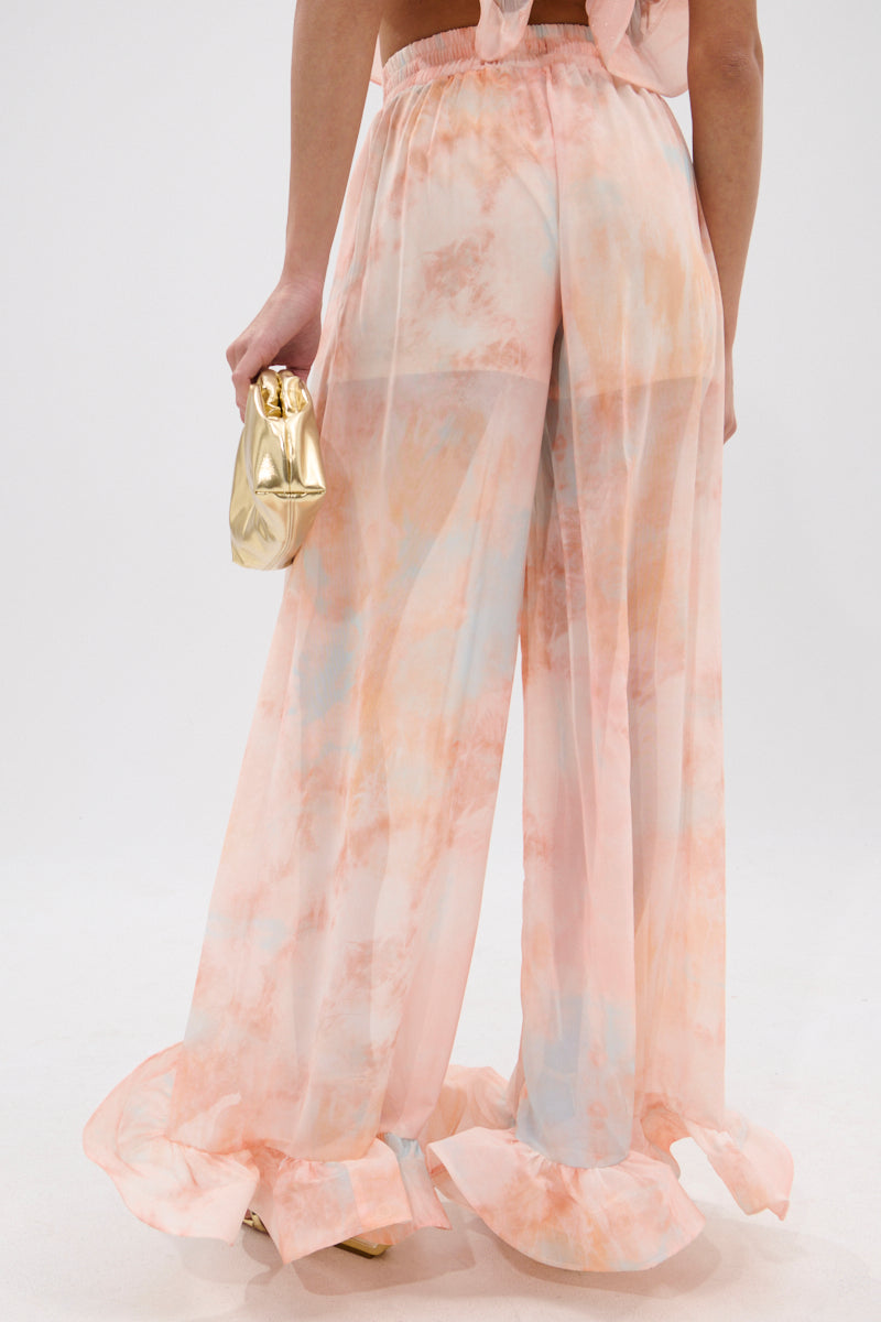 A person faces away wearing the COLORFUL SKY OMBRE PALAZZO PANT, featuring a peach and blue gradient and ruffled hems. They pair it with gold heels and a shiny gold clutch. 4