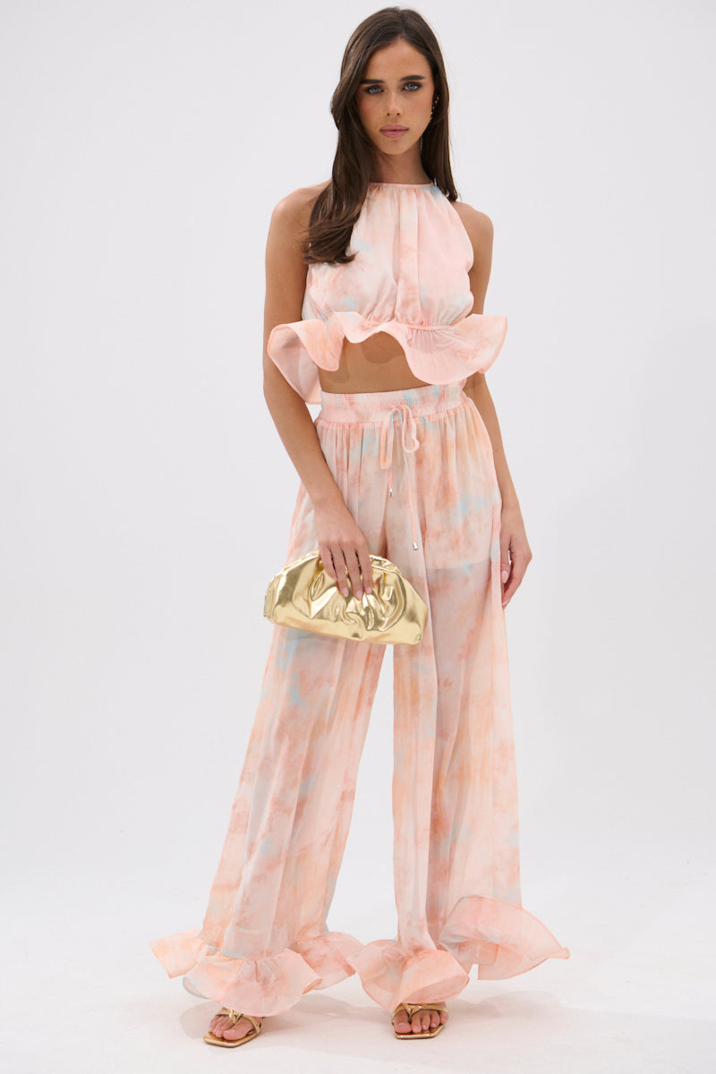 A woman models the COLORFUL SKY OMBRE PALAZZO PANT, a pastel pink and peach sheer two-piece with ruffle details, paired with strappy sandals and a metallic gold clutch against a plain white background. 5
