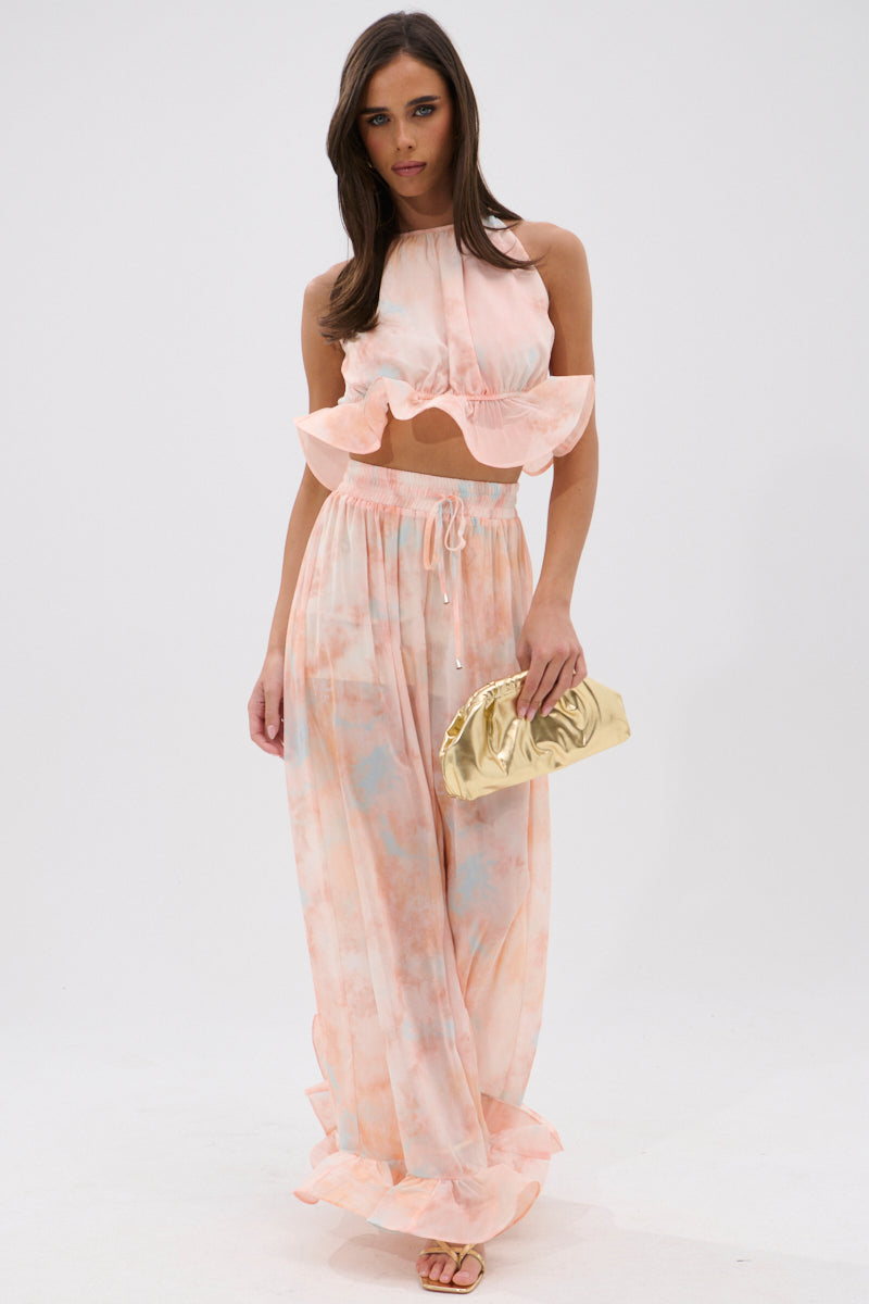 A woman stands against a white background wearing the COLORFUL SKY OMBRE PALAZZO PANT in soft peach and blue, paired with a sleeveless ruffled crop top, metallic gold clutch, and sandals. 6