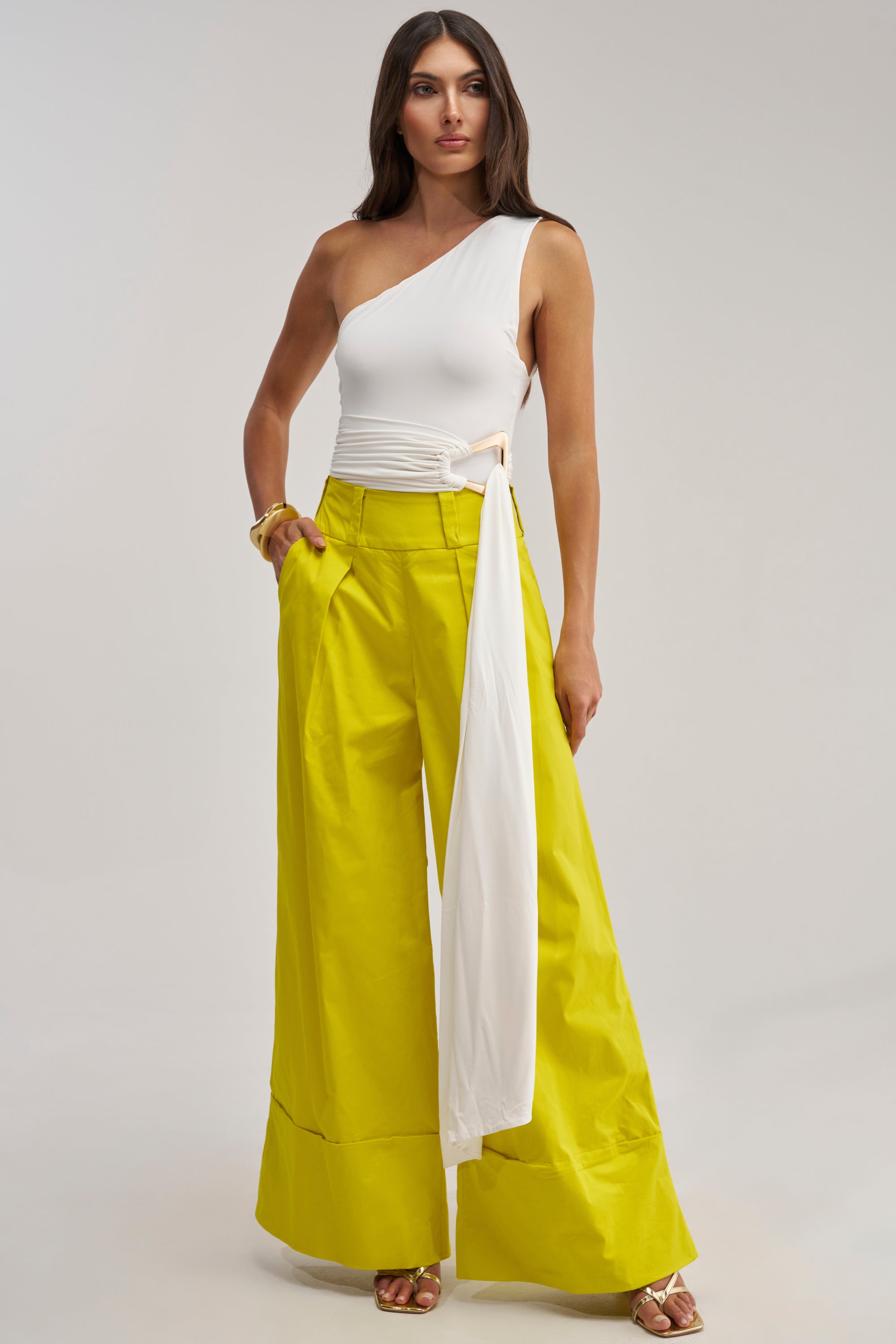 A woman with long brown hair confidently poses in the FEELING GOOD BODYSUIT WITH BELT, a chic white one-shouldered bodysuit featuring a long sash, paired with wide-leg, high-waisted yellow pants—perfect for a date night look.