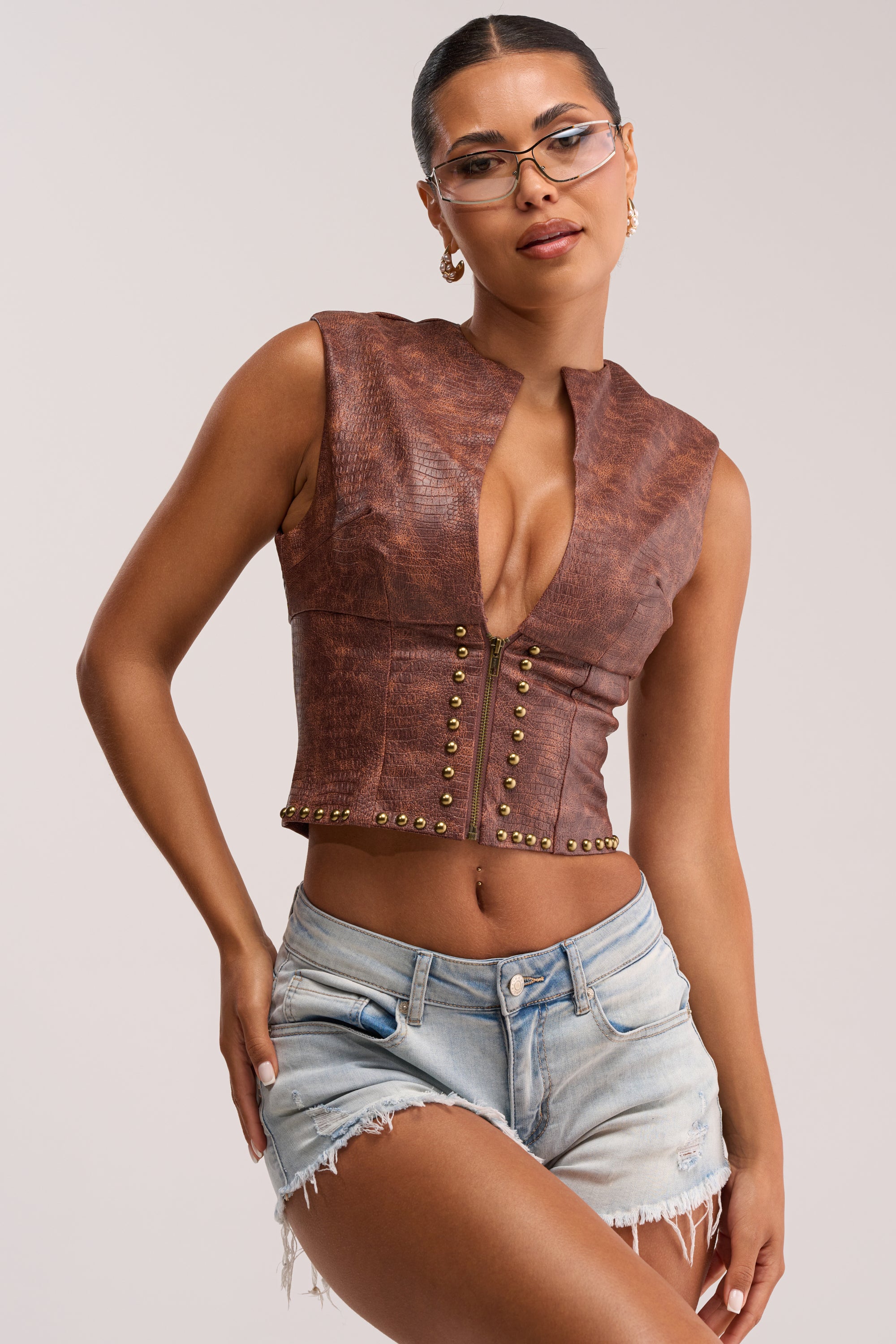 A woman in glasses poses confidently against a plain light background, wearing the DOWN FOR WHATEVER FAUX LEATHER CORSET TOP with a lace-up back and light blue distressed denim shorts.