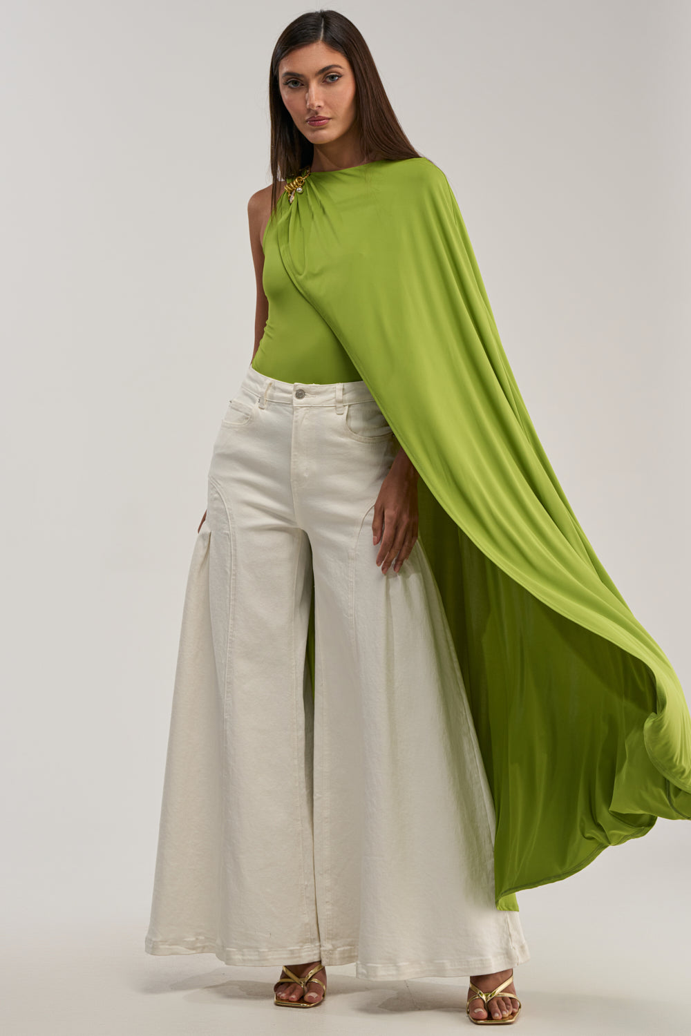 A woman confidently wears the NOT YOURS CAPE BODYSUIT IN CHARTREUSE with high-waisted, wide-leg white pants and gold sandals, creating a bold date night look against a plain background.